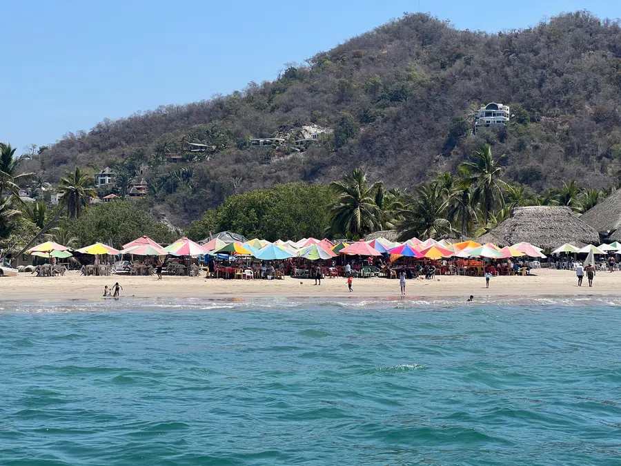 View from the water toward La Manzanilla's palm-lined beach, colorful umbrellas and beachfront restaurants.