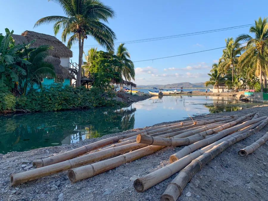 A calm lagoon framed by palms and rustic buildings opens toward the beach and Pacific beyond.