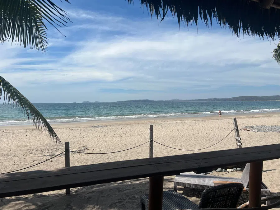 A shaded beachfront table looks across the sand to the calm blue water of La Manzanilla Bay.