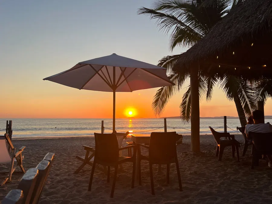 The setting sun glows beside a beach umbrella and palms at the end of a warm La Manzanilla day.