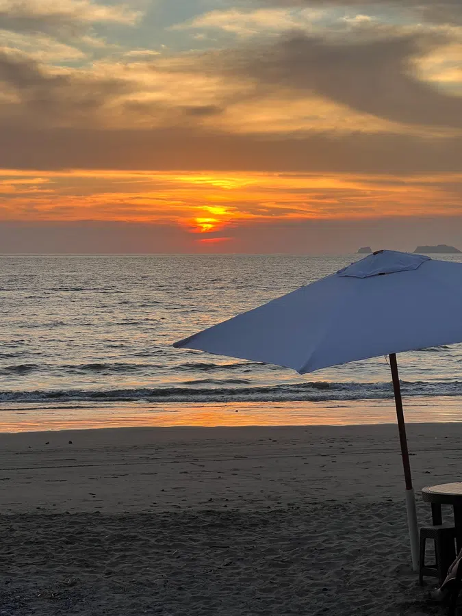 Soft orange sunset light spreads across the Pacific beyond a beach umbrella.
