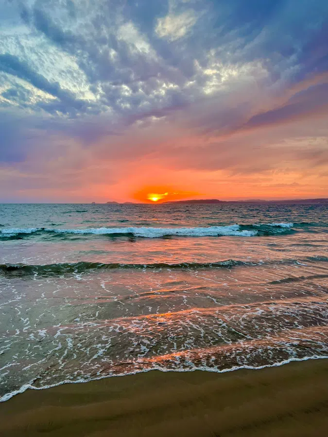 The sun drops toward the horizon as warm color reflects across the breaking waves.