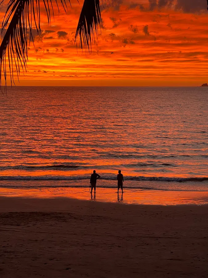 Two figures stand at the water's edge beneath an intensely orange tropical sunset.