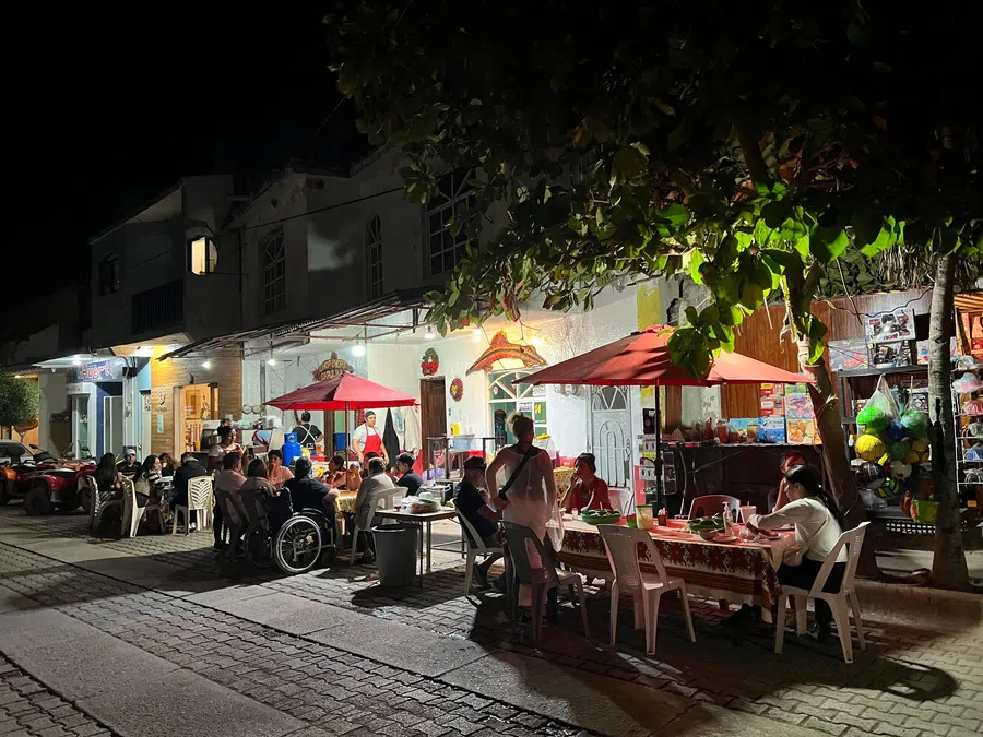 Families and friends gather at casual street-side tables beneath red umbrellas and leafy trees.