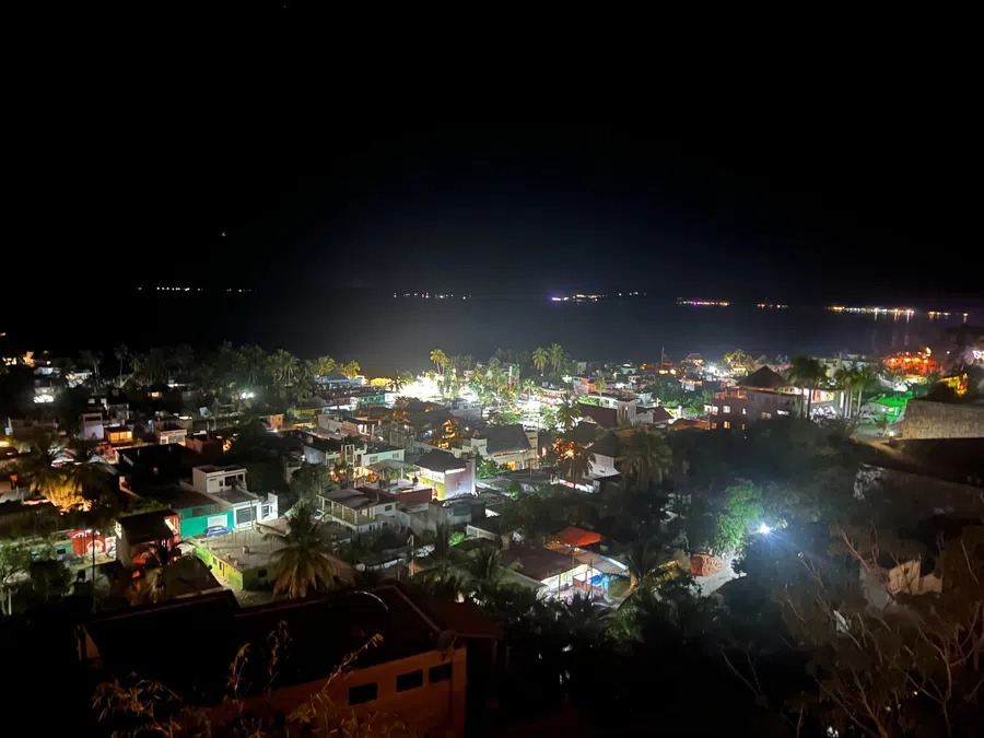 Nighttime hillside view over the illuminated village and the dark Pacific beyond.
