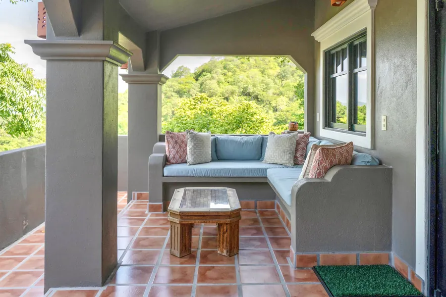 Covered outdoor seating area in front of the four-bedroom residence, with built-in cushioned sectional seating and views toward the lush hillside.