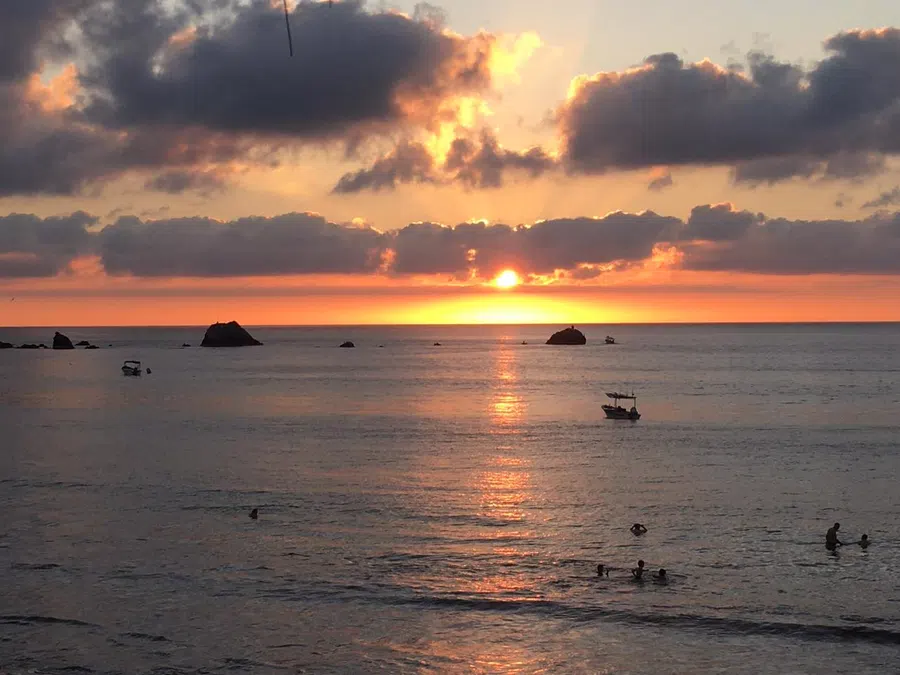 The sun sets between offshore rocks while swimmers and small boats dot the calm evening water.