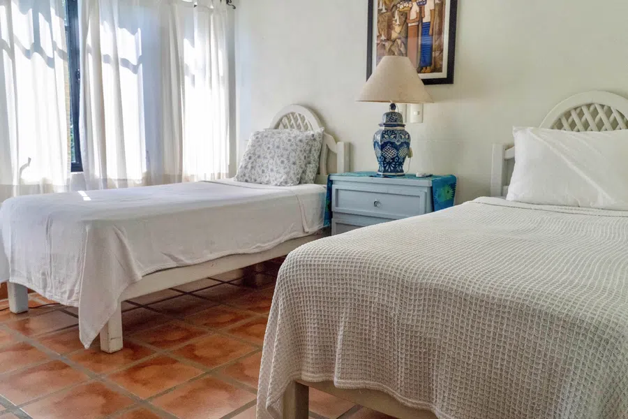 Airy guest bedroom with two beds, white linens and traditional terracotta tile flooring.