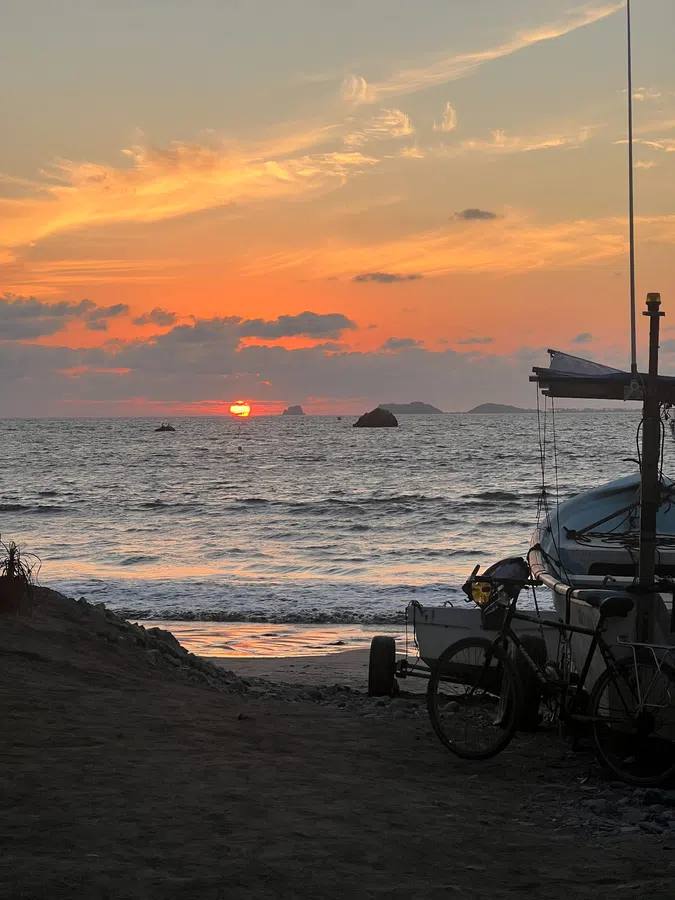 A working beach scene at sunset, with a fishing boat and bicycle silhouetted against the glowing Pacific.