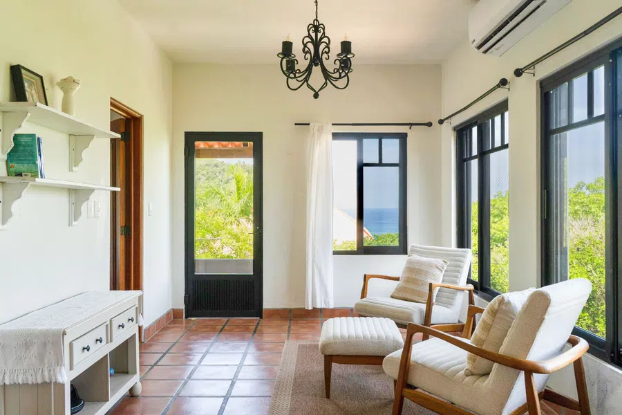 Light-filled sitting room with ocean glimpses, terracotta floors and large black-framed windows.